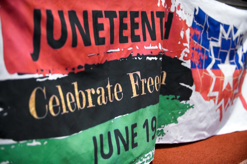 Banner with red, black, and green stripes that fade into a white background. Across the red stripe is the text Juneteenth. Below that is the Black stripe with text Celebrate Freedom. Below that is the green stripe with the text June 19. Across from the stripes is a map of the United States with half of the map blue and half red. A white 5-point star is in the middle of the map.